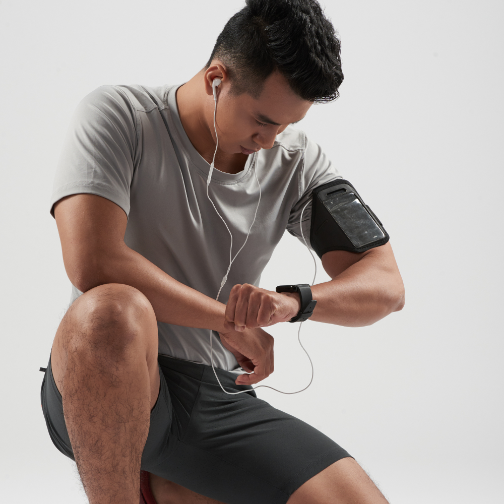Man in athletic wear with earphones and arm sleeve, checking time on wristwatch against a plain background

