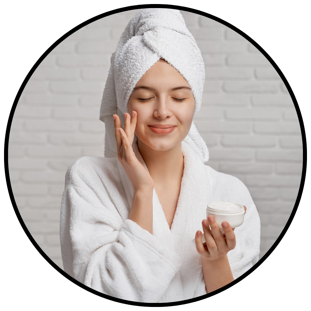 Woman in a white robe with a towel on her head, holding a bowl and applying cream to her face.

